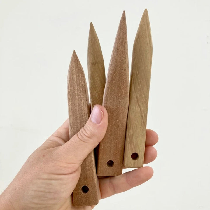 A hand holding four pointed wooden stakes or markers with pre-drilled holes, shown against a plain off-white background.