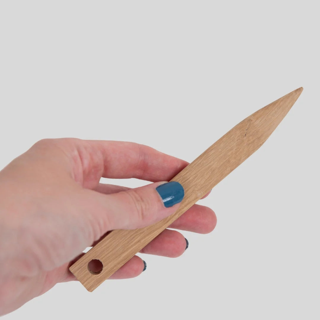 A hand with blue nail polish holds a light natural wood stake or tool against a plain light gray background.