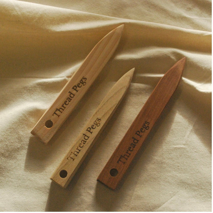 Three wooden thread pegs in different wood tones, engraved with the text 'Thread Pegs' and featuring a hole at one end, arranged on a draped beige fabric background.