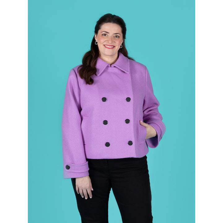 A woman modeling a lavender purple double-breasted jacket with black buttons, paired with black pants, against a solid turquoise background.