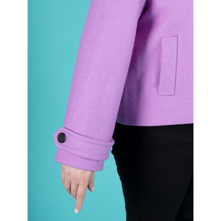 Close-up of a person wearing a lavender-purple coat with a buttoned cuff and black pants, standing against a solid teal background.