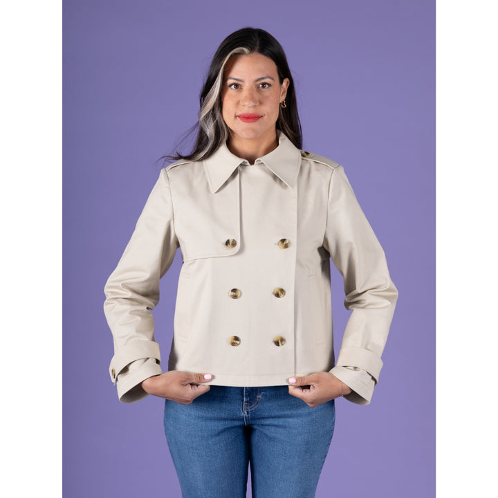 A woman modeling a light beige double-breasted trench jacket with gold buttons, paired with blue jeans, against a solid purple background.