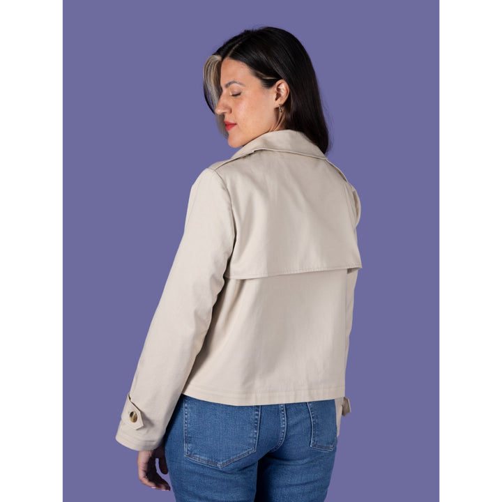 A woman shown from the back wearing a light beige trench-style jacket and blue jeans, standing against a solid purple background.
