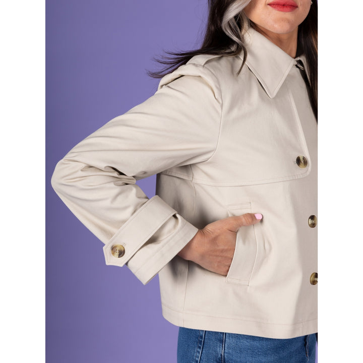 A woman wearing a light beige trench-style jacket with gold-tone buttons and a pointed collar, paired with blue jeans, set against a solid purple background.