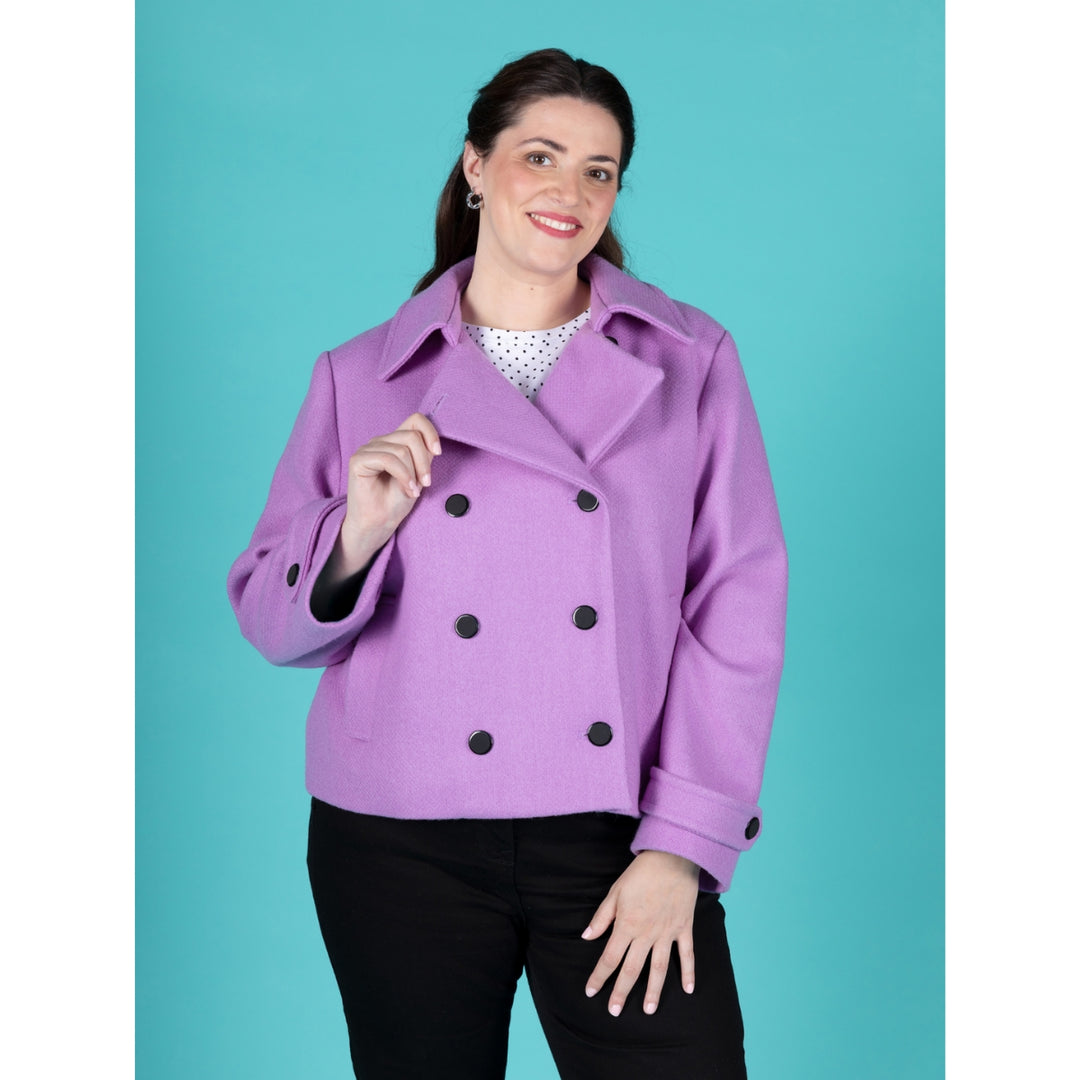 A woman modeling a lavender-purple double-breasted peplum-style coat with black buttons, paired with a white polka-dot top and black pants, set against a solid teal background.