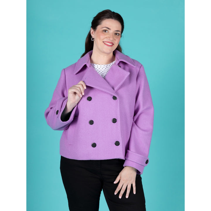 A woman modeling a lavender-purple double-breasted peplum-style coat with black buttons, paired with a white polka-dot top and black pants, set against a solid teal background.
