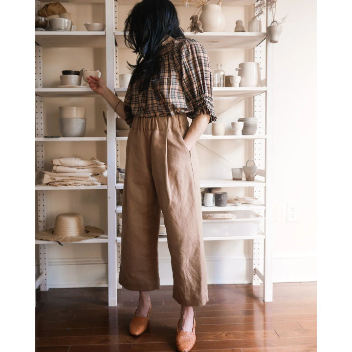 Person in plaid shirt and brown pants standing in front of a white shelving unit with ceramic items.