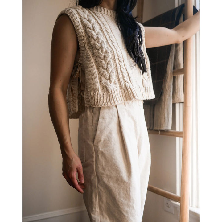 Person wearing a beige knitted vest and white pants standing near a wooden ladder.