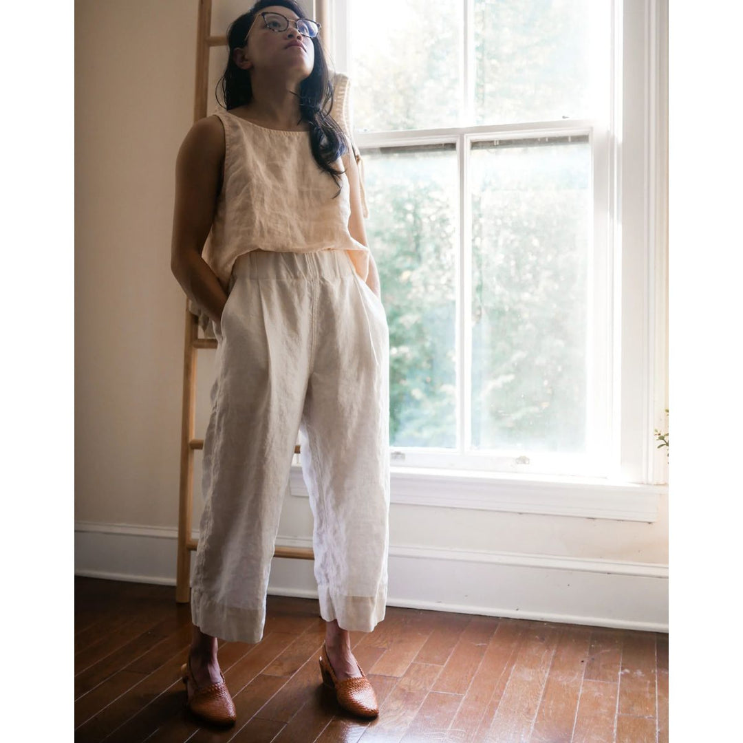 Woman standing in a room with a large window, wearing a beige top and white pants.