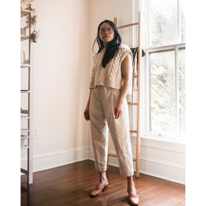 Woman wearing a beige knit top and pants standing in a room with wooden flooring and a window.