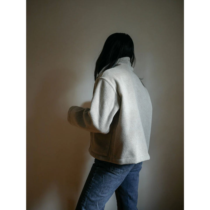 Person wearing a light gray sweater and blue jeans against a plain wall.
