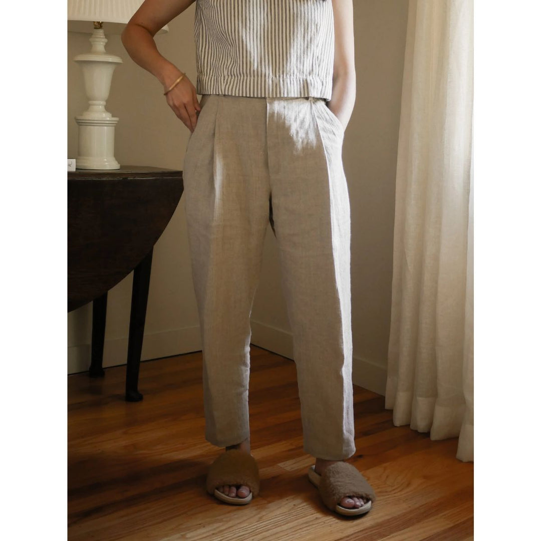 Person wearing beige pants and a striped top standing in a room with wooden floor and white curtain.