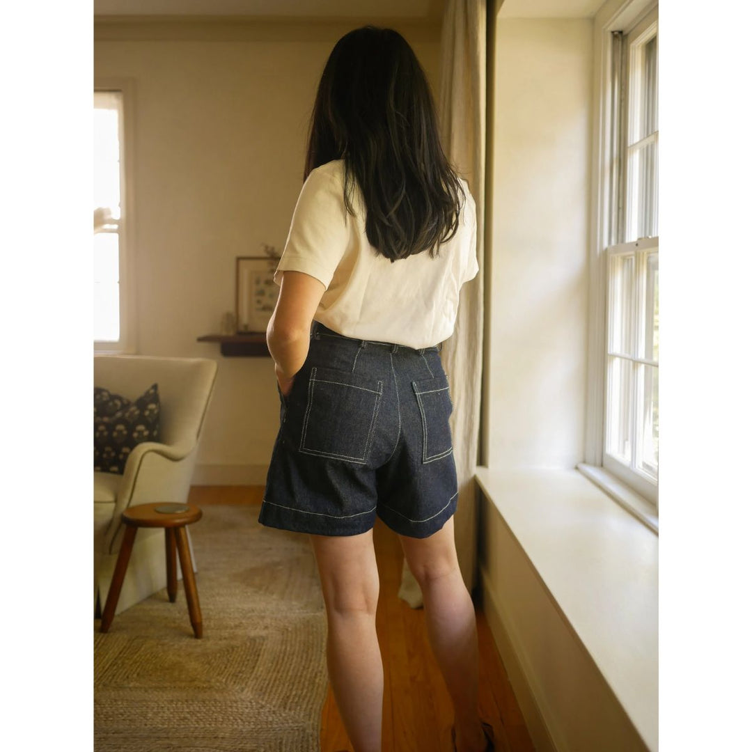 Person wearing a white shirt and dark shorts standing in a room with a window and furniture.