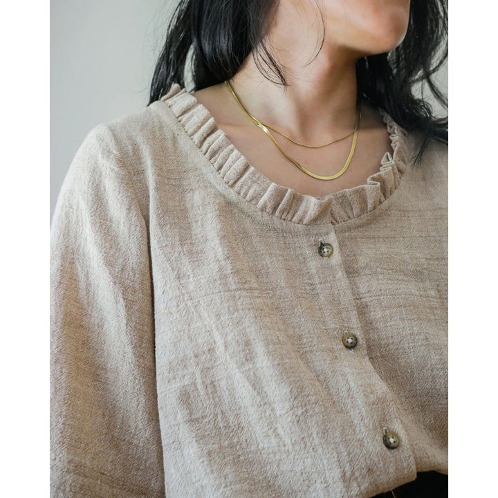 Person wearing a beige textured blouse with gold buttons and layered necklaces.