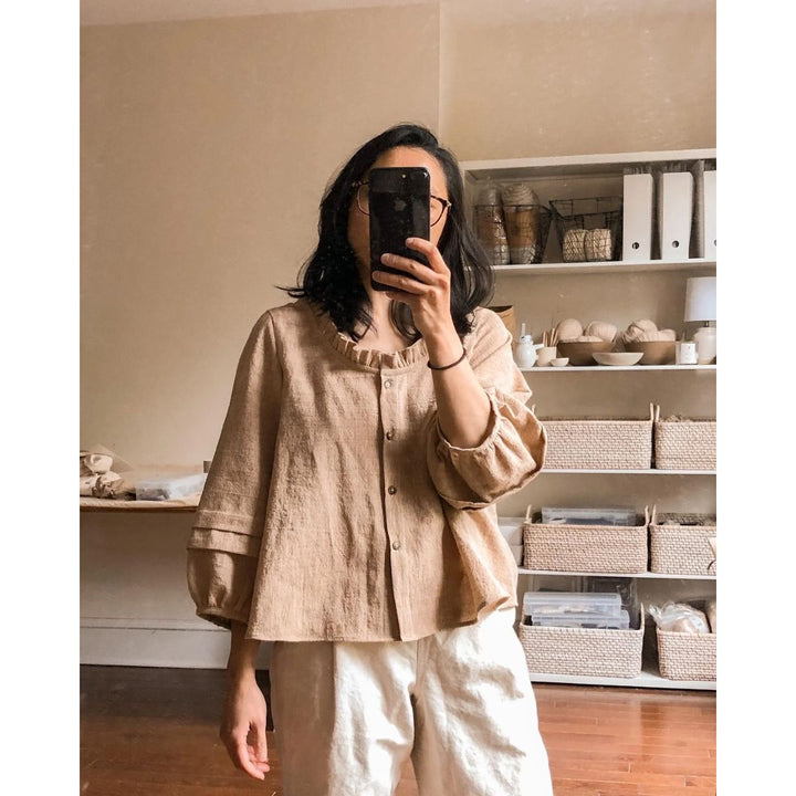 Person wearing a beige shirt and white pants taking a mirror selfie in a room with shelves and storage baskets.
