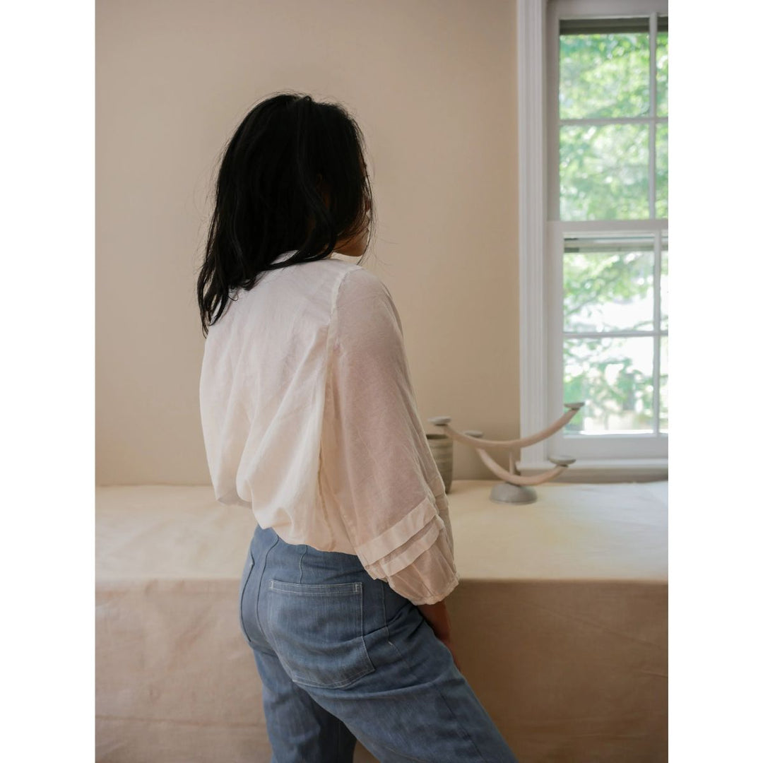 Person wearing a white blouse and blue jeans standing in a room with a window and decorative object.