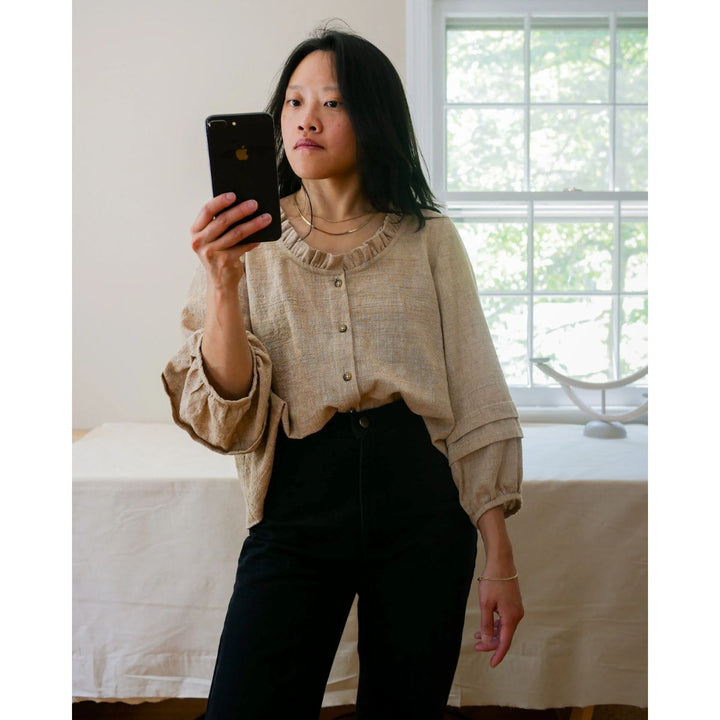 Person taking a mirror selfie wearing a beige blouse and black pants in a room with a window.