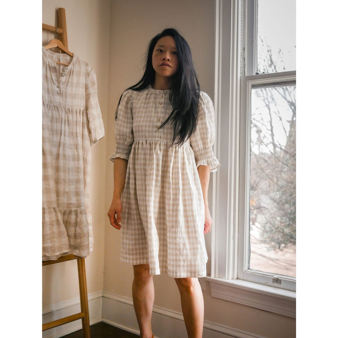 Woman wearing a checkered dress standing next to a window in a room with another dress on a rack.
