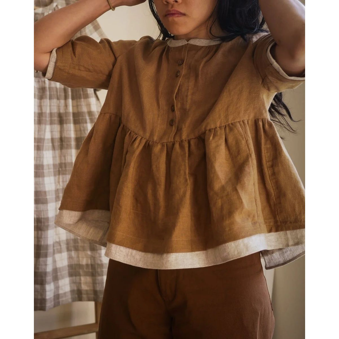 Person wearing a brown top and pants with a plaid curtain in the background