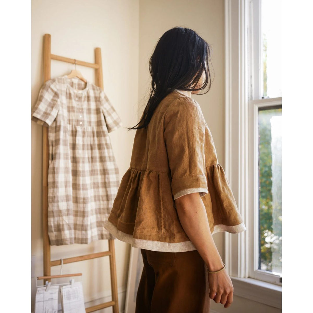 Woman in a brown dress standing next to a rack with a plaid dress in a room with a window.