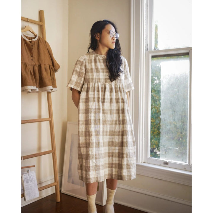 Woman in a plaid dress standing by a window in a room with a wooden rack and framed picture.