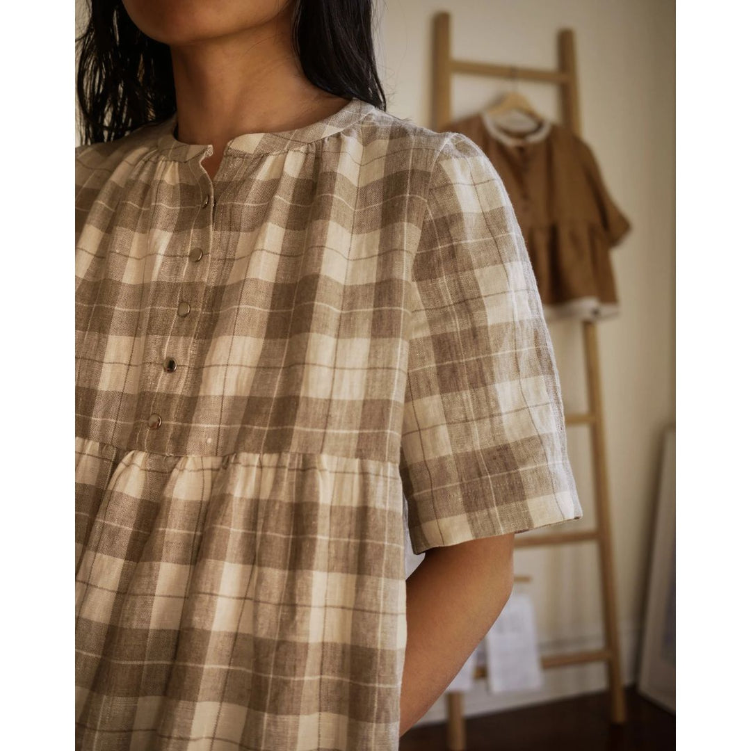 Person wearing a brown and beige plaid dress in a room with a wooden ladder and coat in the background.