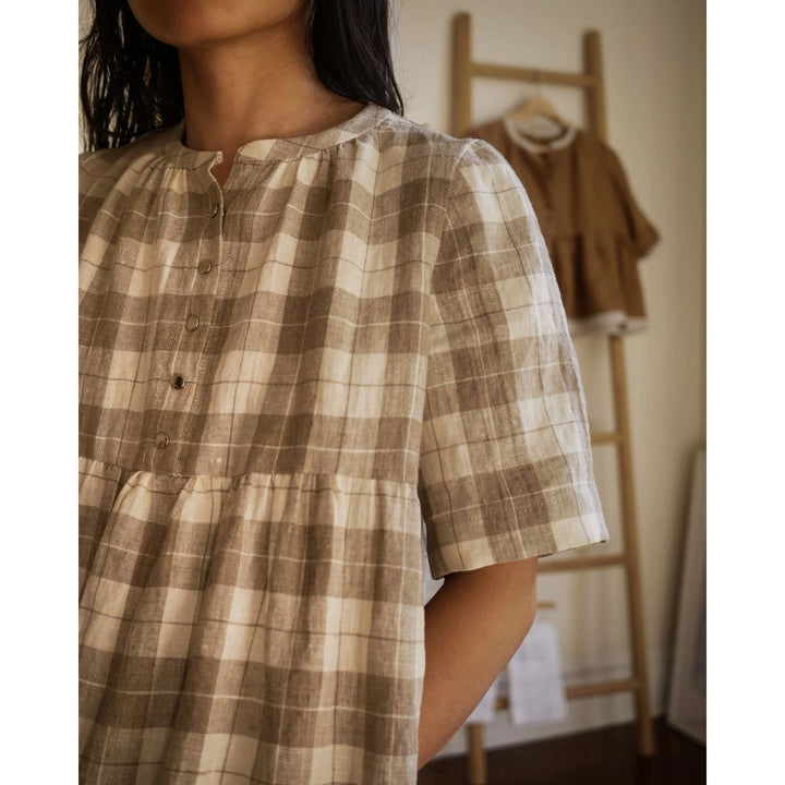 Person wearing a brown and beige plaid dress in a room with a wooden ladder and coat in the background.