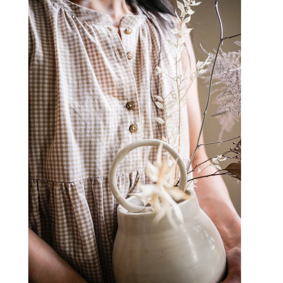 Person holding a white ceramic teapot with dried plants, wearing a checkered dress.