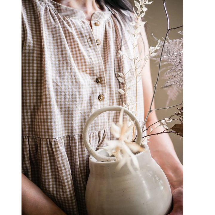 Person holding a white ceramic teapot with dried plants, wearing a checkered dress.