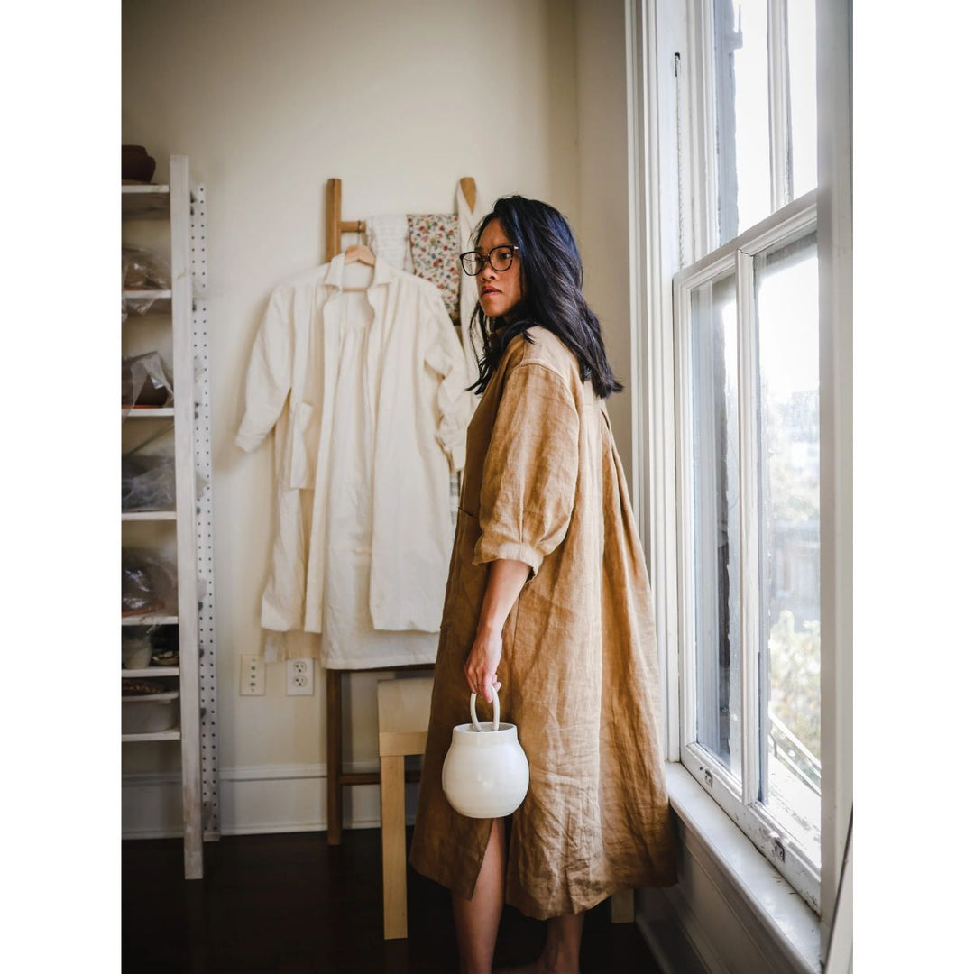 Woman in a beige dress holding a white bag in a room with a window and clothing rack.