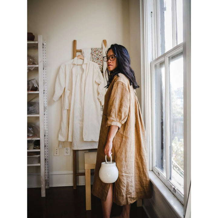 Woman in a beige dress holding a white bag in a room with a window and clothing rack.
